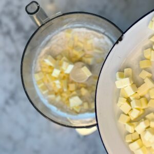Overhead view of chilled butter cubes being added to the bowl of a food processor filled with pie crust dry ingredients.